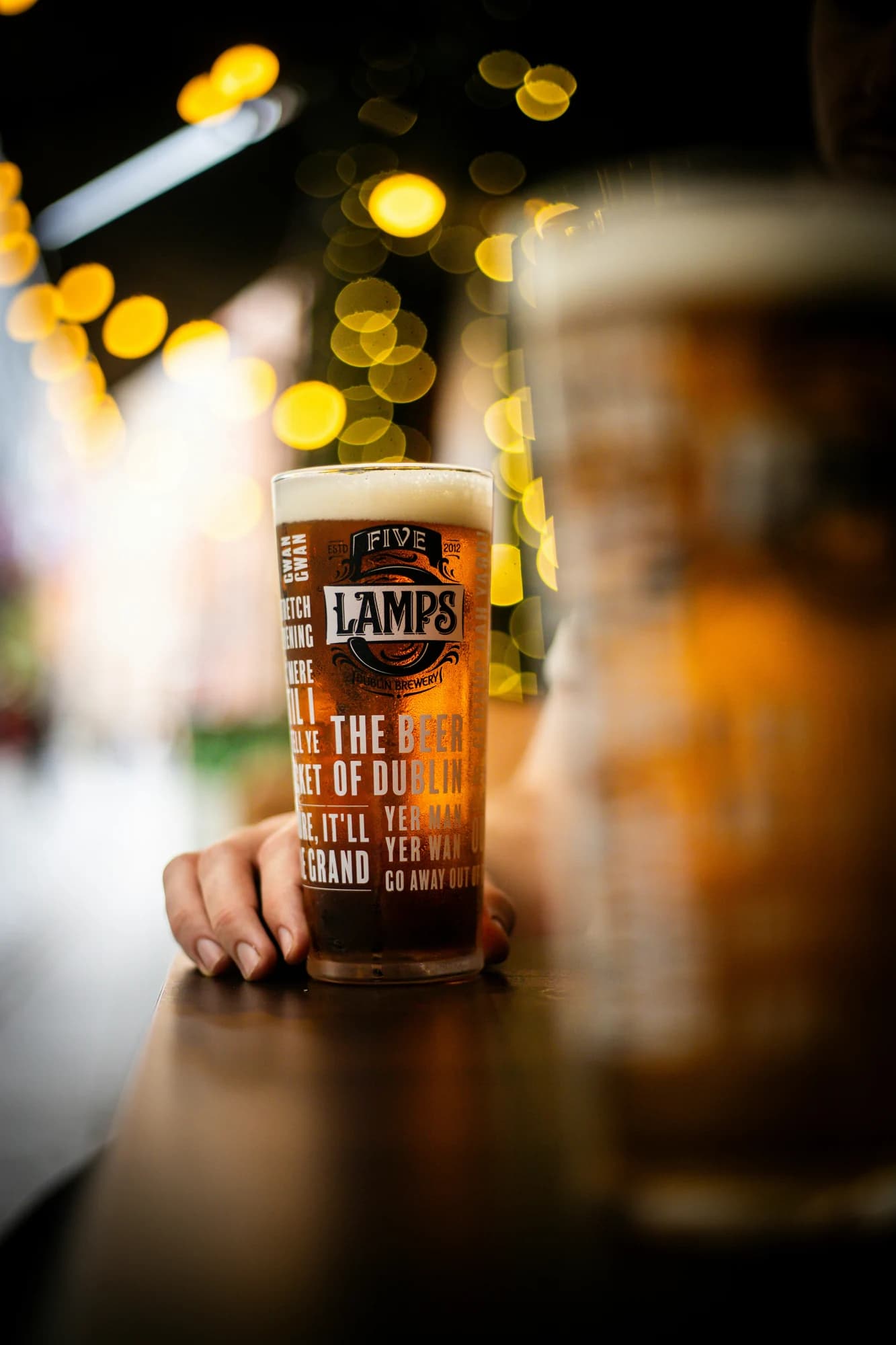 Hand holding a pint of beer with bokeh lights — representative photo, not taken at The Arlington Bar